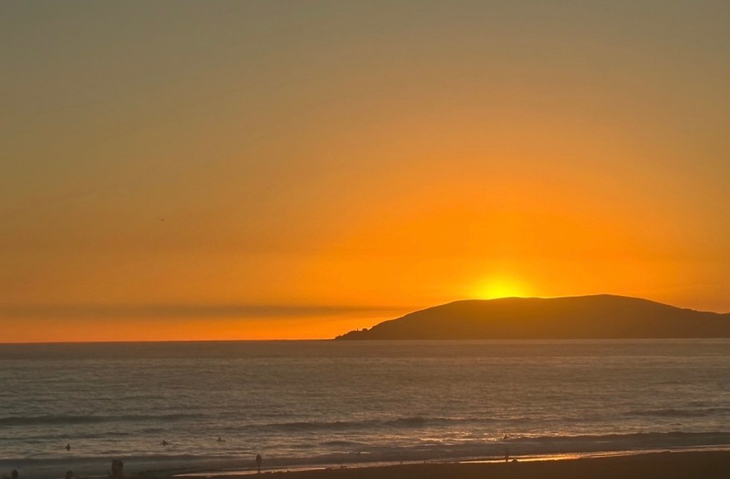 Golden-orange sunset over the Pacific Ocean, with sun disappearing behind a coastal hillside
