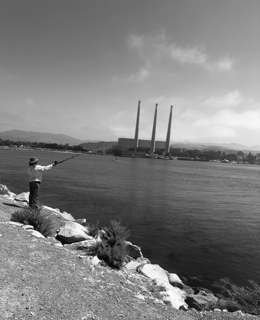 Black-and-white photograph of a person fishing across Morro Bay with three power plant smokestacks in the distance.